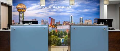 A hotel reception area with a cityscape mural, a reception desk, and a view of a pool and buildings.