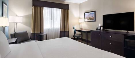 Habitación de hotel con una cama grande, un escritorio, una silla, un televisor y una ventana con cortinas.