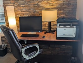 Escritorio de madera con monitor de computadora, teclado e impresora. Una silla de cuero negro está ubicada frente al escritorio.