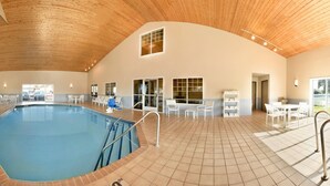 Ein großes Hallenbad mit Fliesenboden, umgeben von weißen Stühlen und Tischen, und einer Holzbalkendecke.
