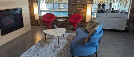 A modern living room with a fireplace, a blue sofa, two red chairs, a white coffee table, and a stone wall.
