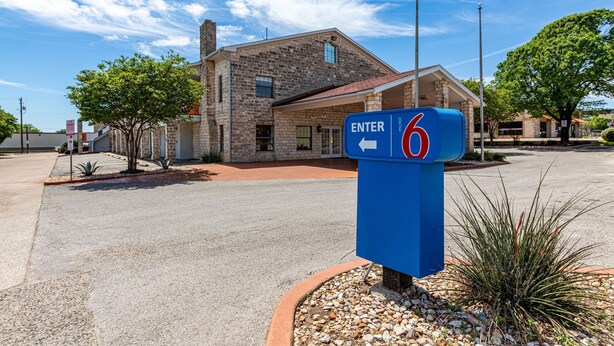 A motel with a stone facade and a red roof, a blue sign indicating room number 6, and a clear blue sky.