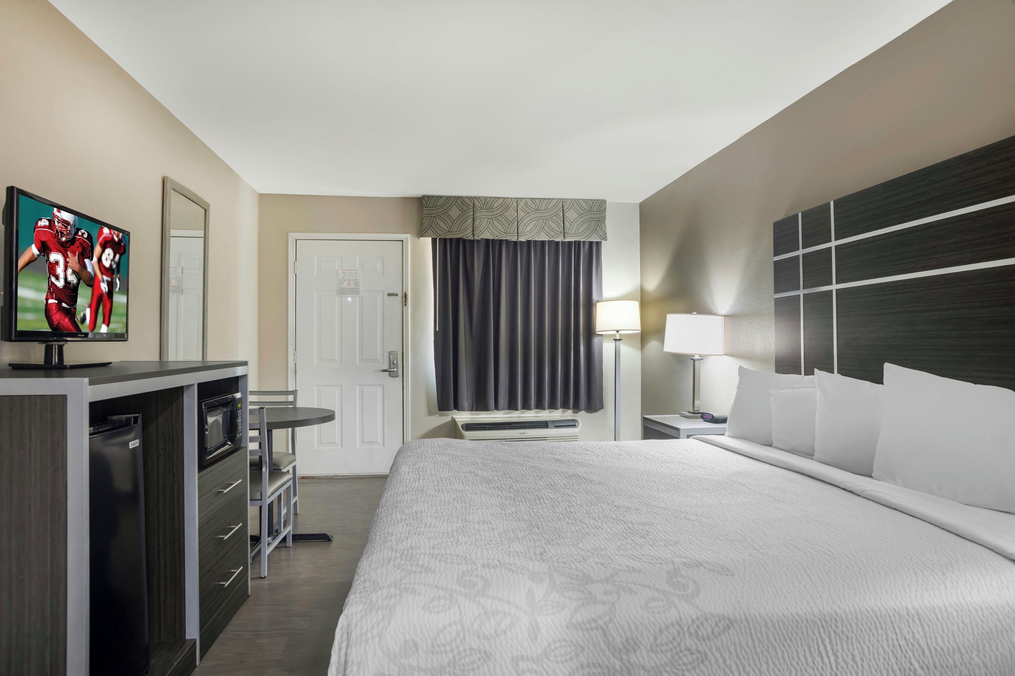 Ein Hotelzimmer mit einem Bett, einem Schreibtisch mit Mikrowelle, einem Fernseher, einer Tür, einem Fenster mit Vorhängen und einem Nachttisch mit Lampe.