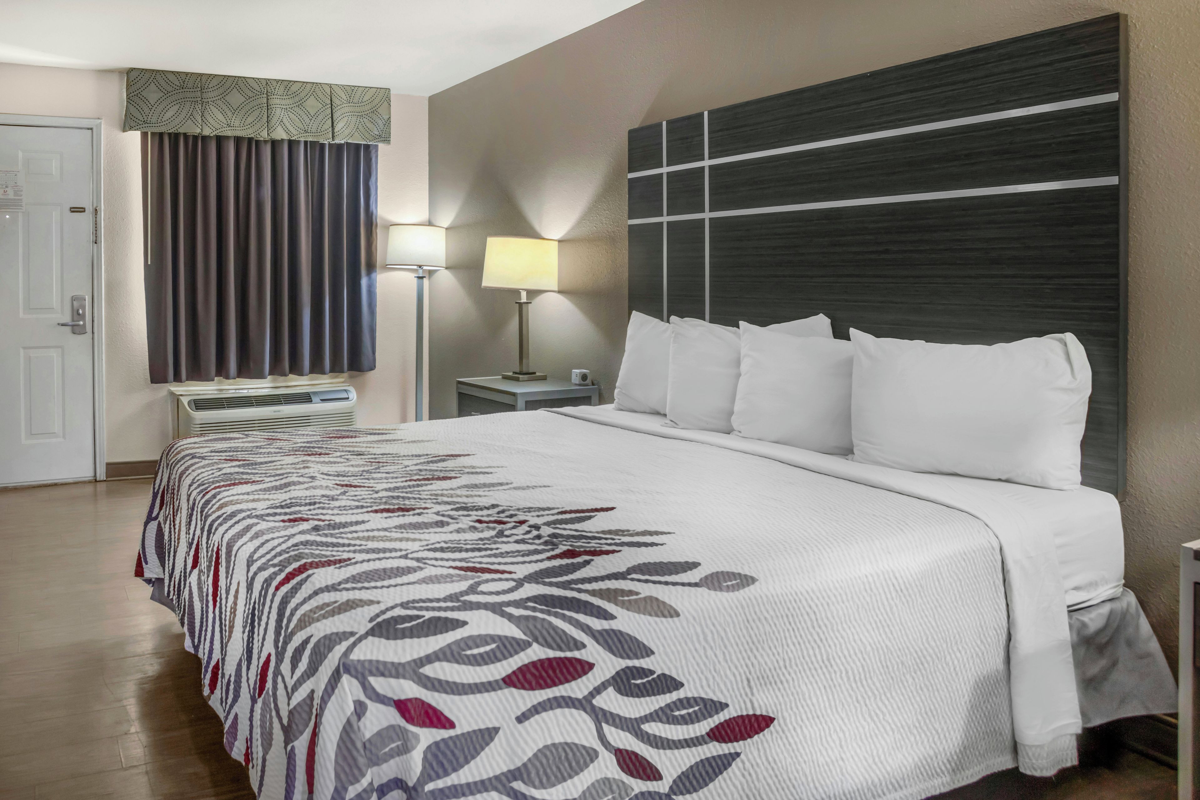 A hotel room with a bed, a nightstand, a lamp, a window with curtains, and a door.