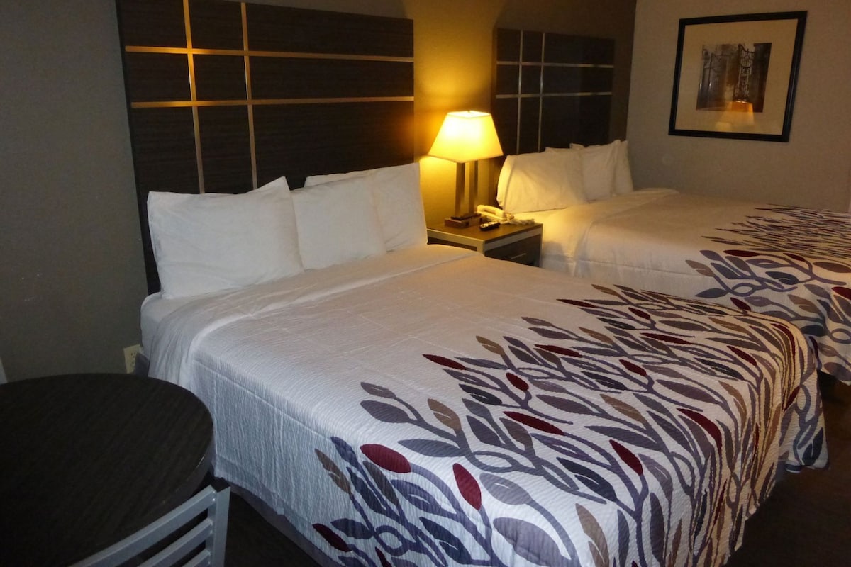 A hotel room with two beds, a nightstand with a lamp, a small table, and a framed picture on the wall.
