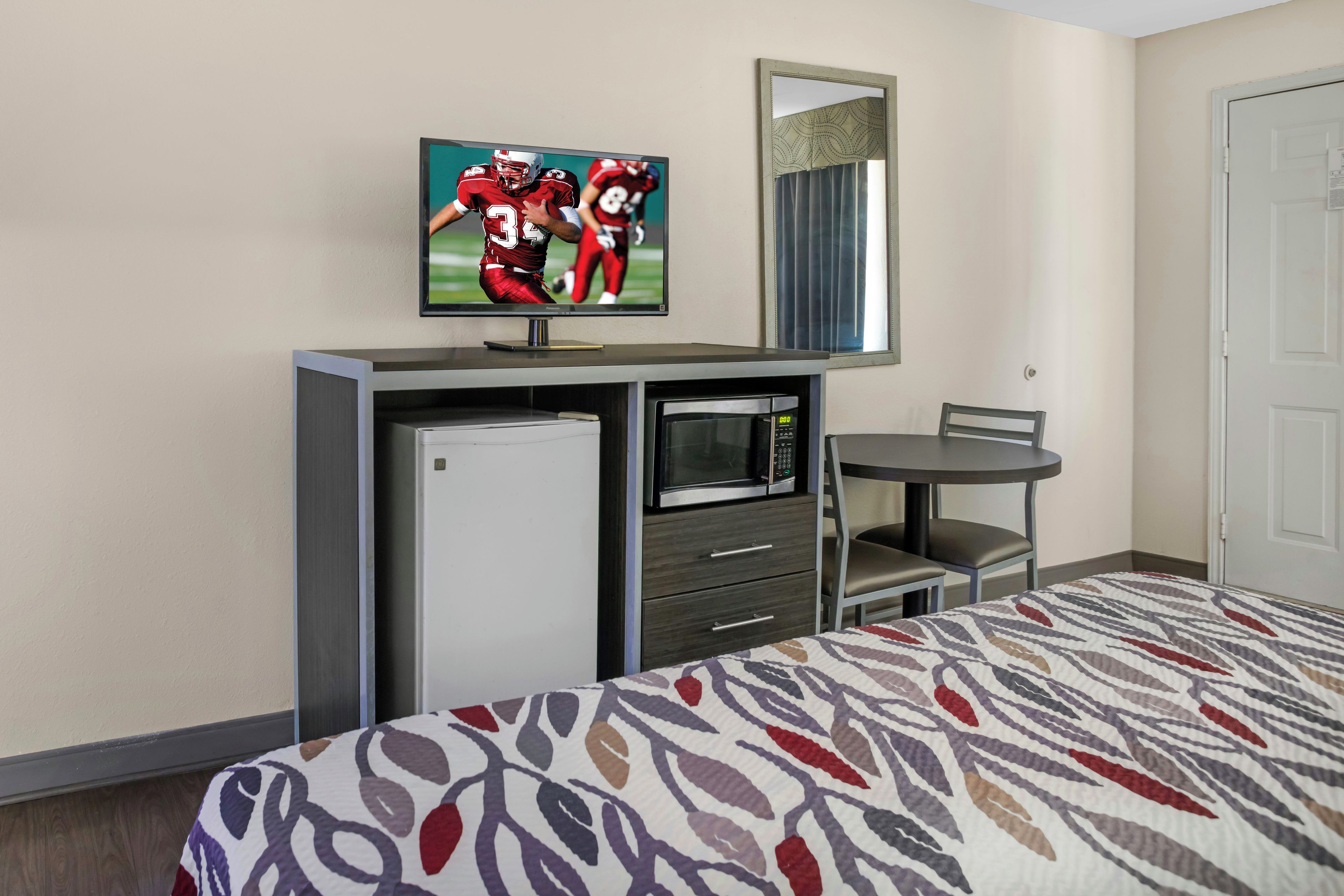 A hotel room with a bed, a television mounted on a dresser, a microwave, a small table with a chair, and a door.