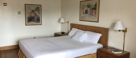 A neatly made bed with white linens and a wooden headboard, flanked by two bedside tables with lamps and a small bedside drawer.