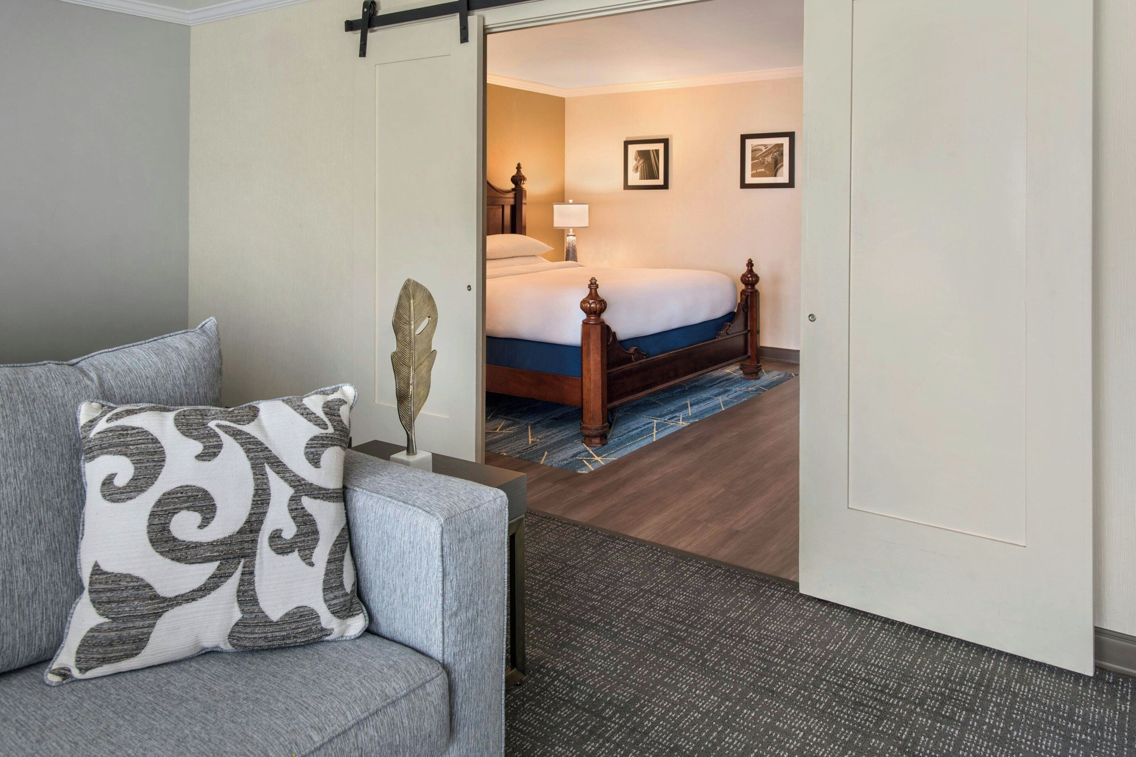 A hotel room with a grey sofa, a decorative leaf, a bed with a wooden headboard, a blue carpet, and framed pictures on the wall.