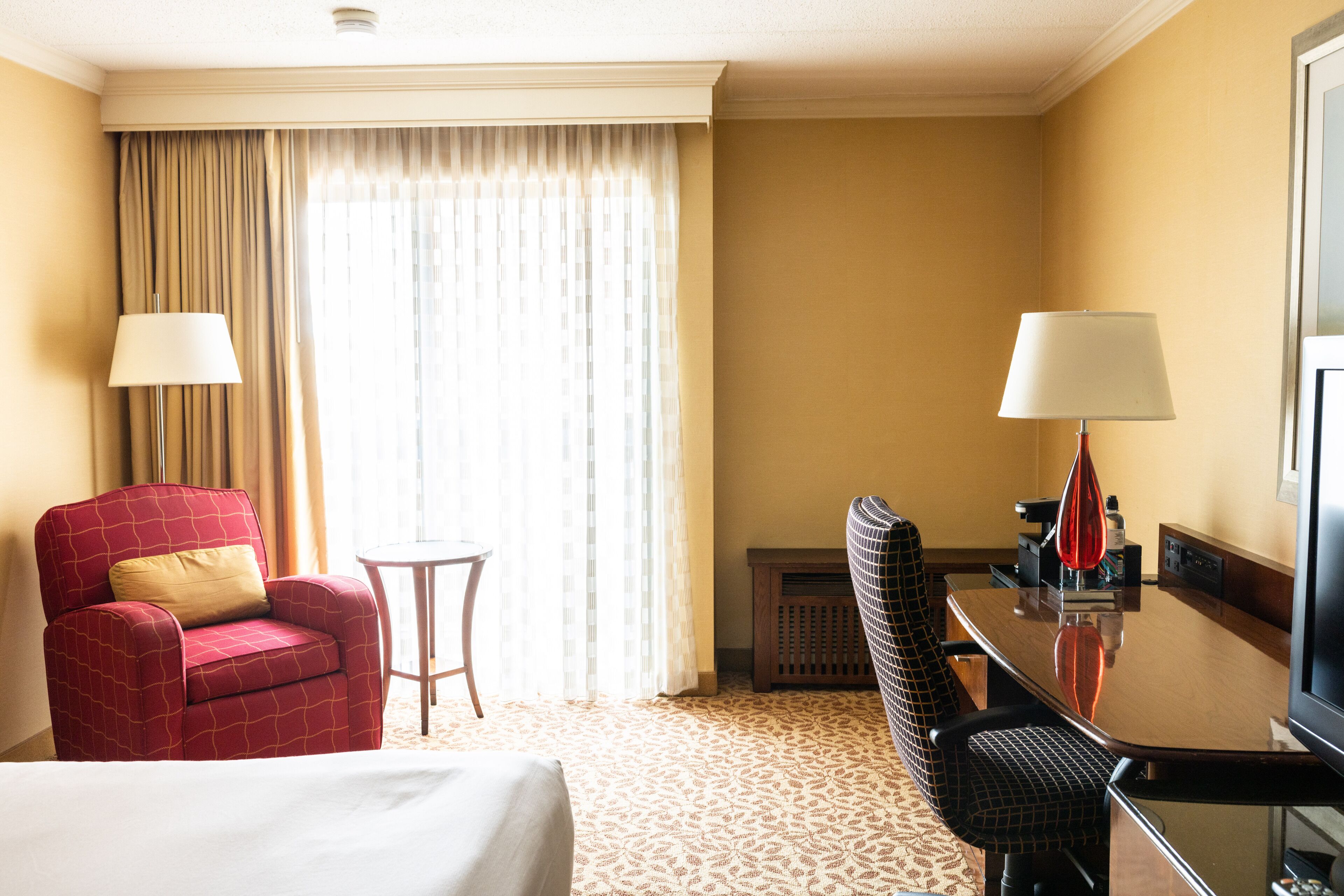 A hotel room with a bed, a red armchair, a small round table, a black office chair, a desk with a lamp, and a television.