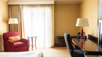 A hotel room with a bed, a red armchair, a small round table, a black office chair, a desk with a lamp, and a television.