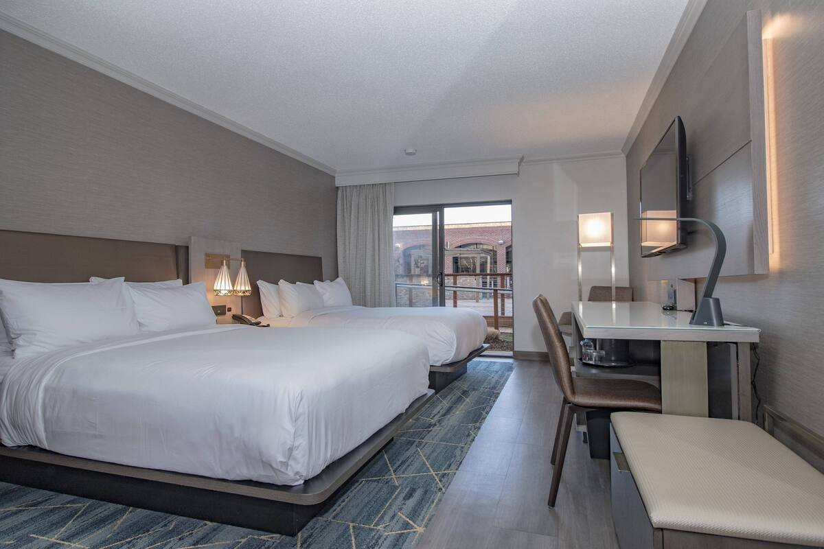 A hotel room with two beds, a desk, a chair, and a large window.
