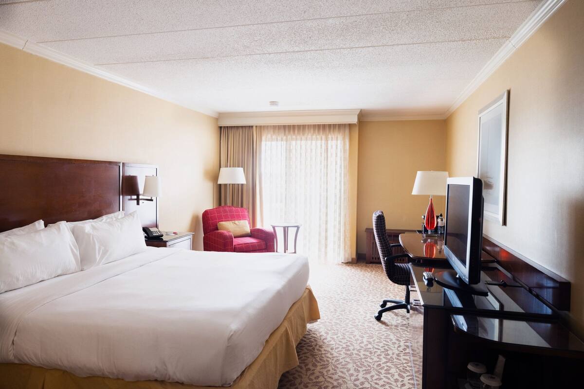 A hotel room with a large bed, a desk with a computer, a chair, a TV, and a window with curtains.