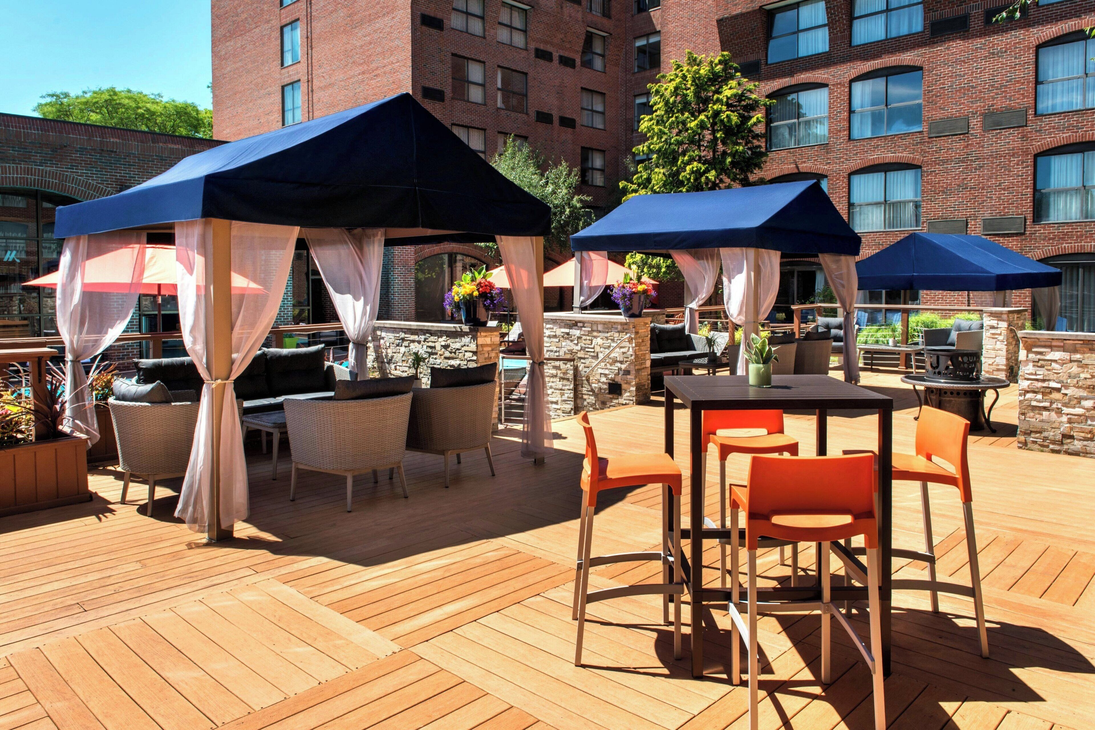 An outdoor patio with wooden flooring, outdoor seating with cushions, and umbrellas providing shade.