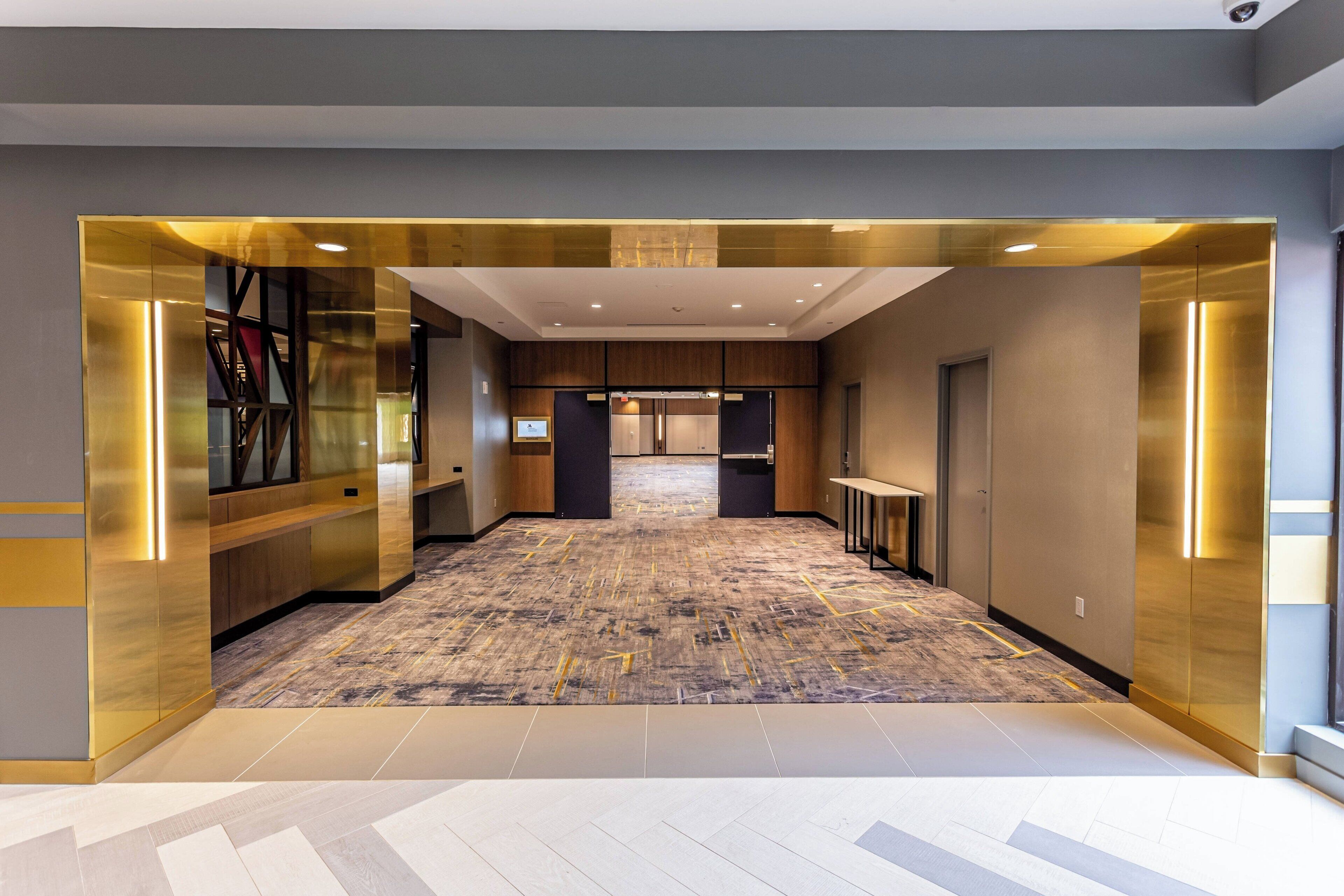 A modern hotel lobby with a central elevator, a patterned carpet, and a glass partition on the left.
