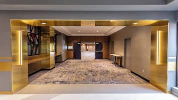 A modern hotel lobby with a central elevator, a patterned carpet, and a glass partition on the left.