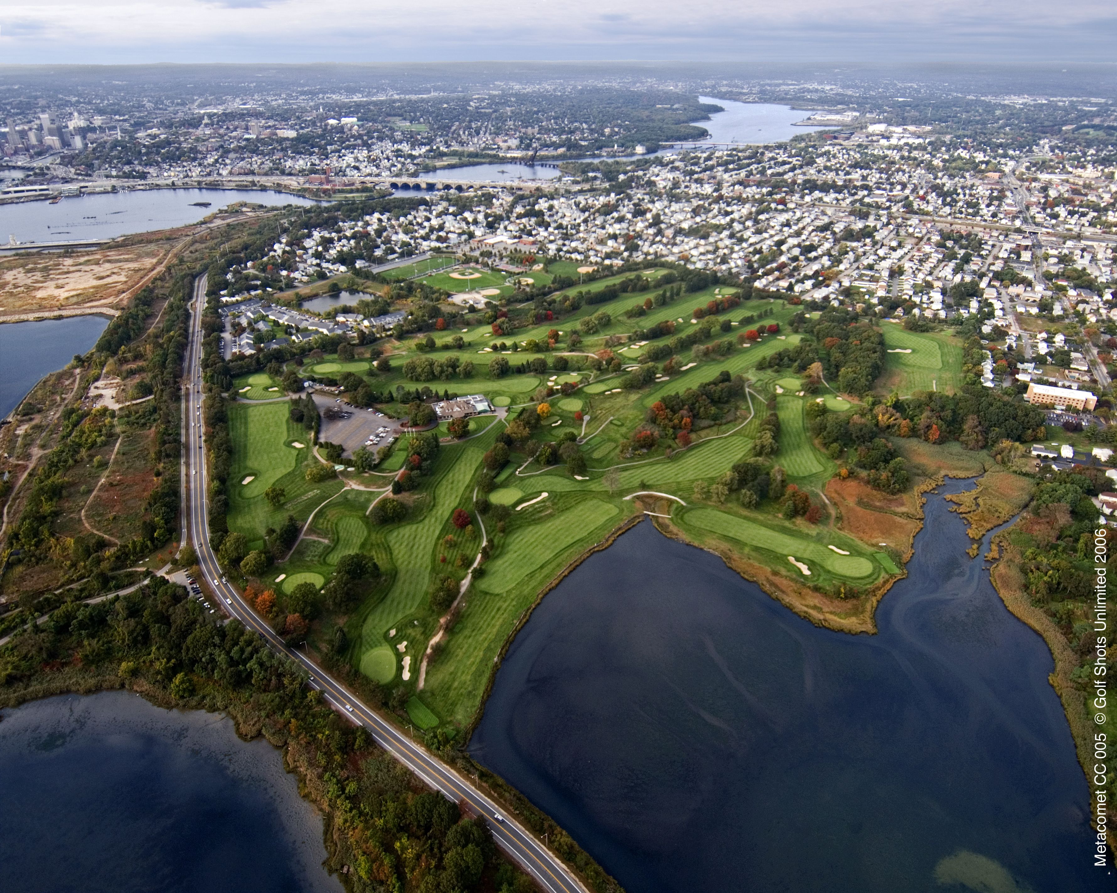 An aerial view of a golf course with a bridge, surrounded by residential areas and a large body of water.
