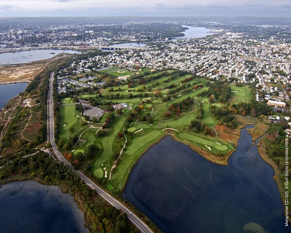 An aerial view of a golf course with a bridge, surrounded by residential areas and a large body of water.