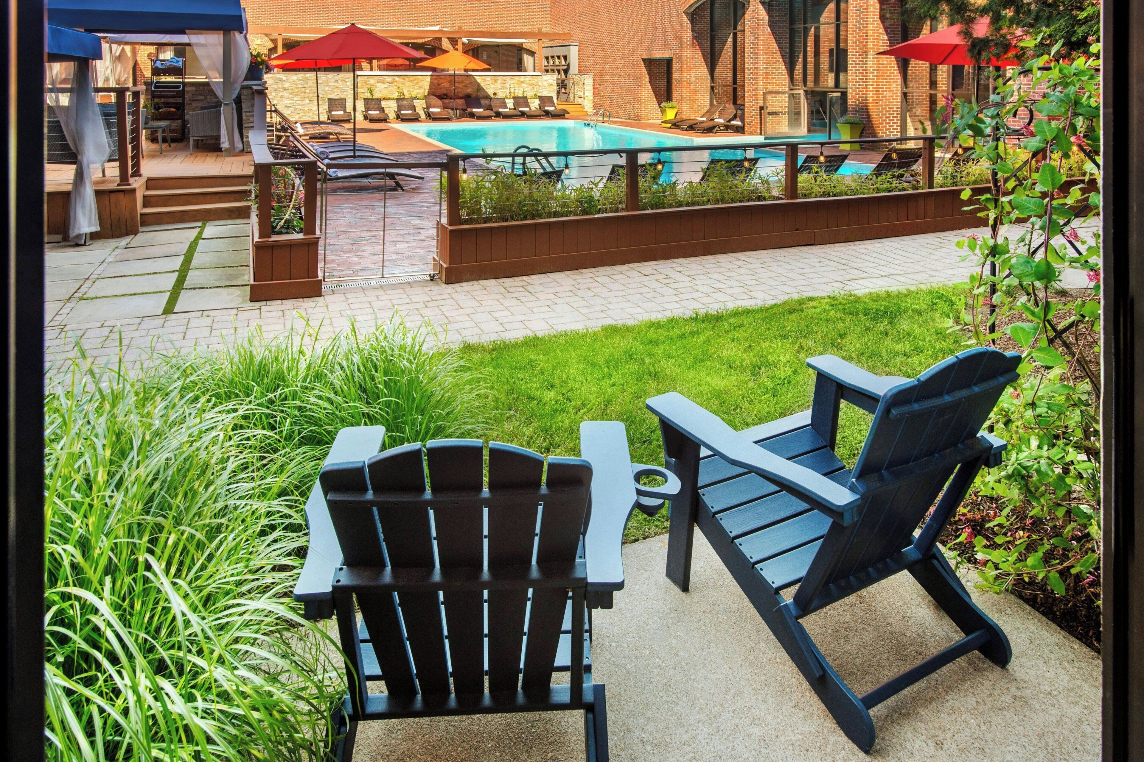 A poolside area with lounge chairs, a pool, and a patio with a brick wall and greenery.