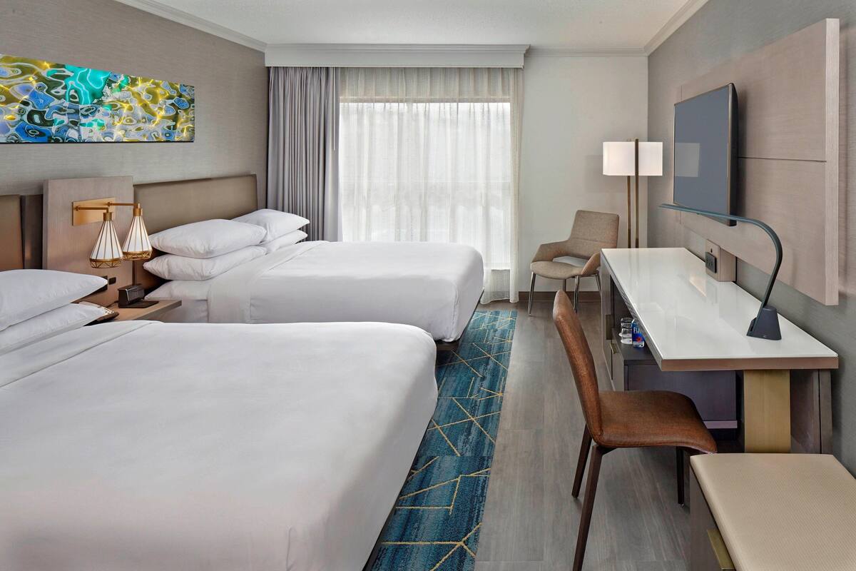A hotel room with two beds, a desk, a chair, a TV, and a large window with curtains.