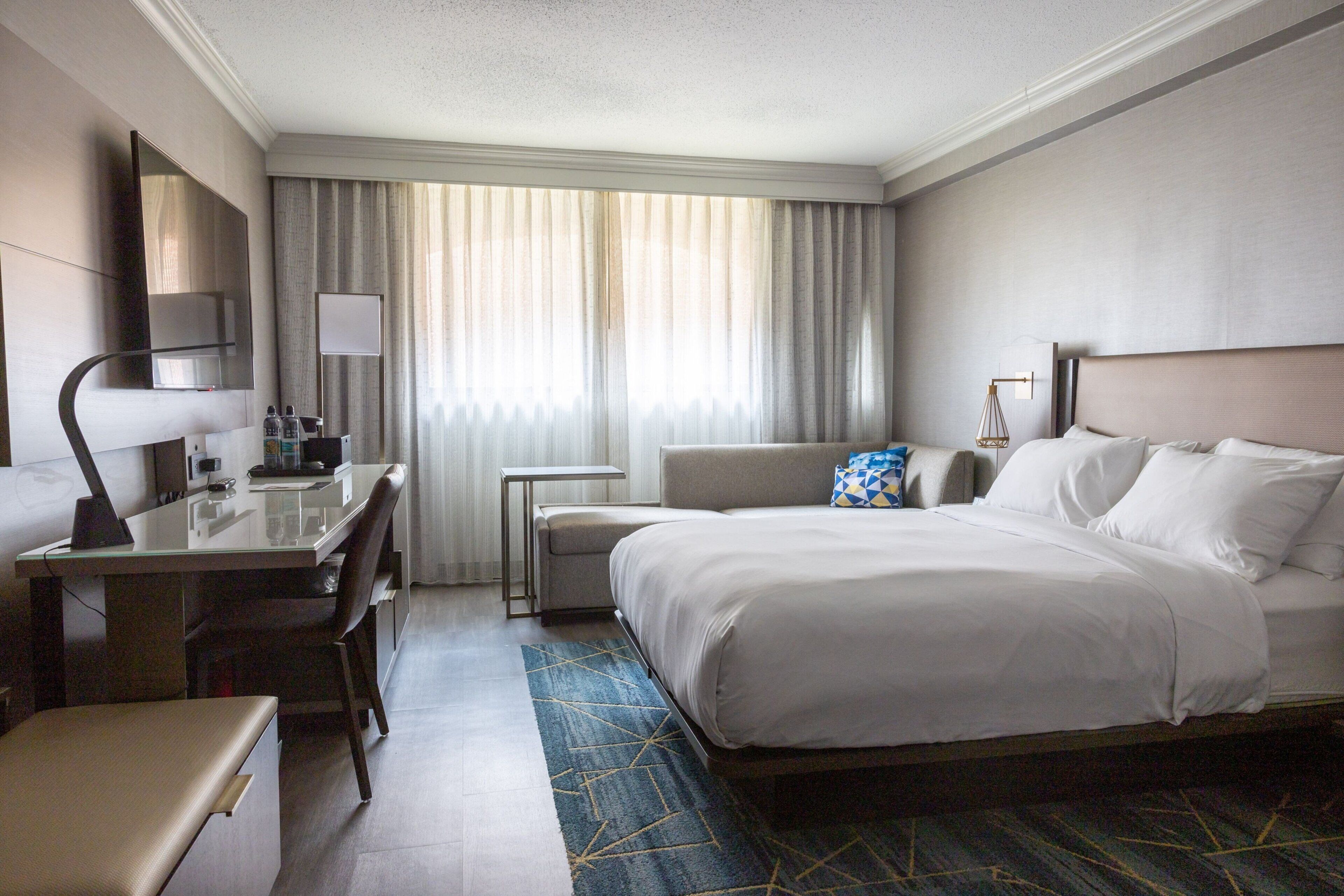 A hotel room with a bed, a desk, a chair, a TV, and a window with curtains.