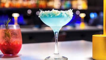 A bar setting with a blue cocktail garnished with a white rim, a red drink with a rosemary sprig, and a yellow candle.