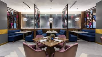 A modern restaurant with a central dining table surrounded by pink chairs, blue booths, and a geometric wall design.