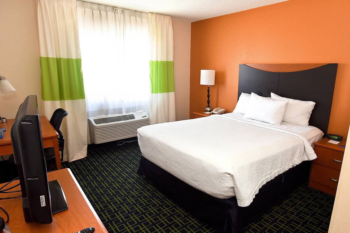Quarto de hotel com cama, escrivaninha com computador, luminária e janela com cortinas.