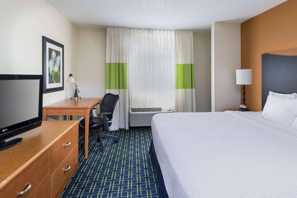 Quarto de hotel com cama, escrivaninha com cadeira, televisão, abajur e um quadro na parede.