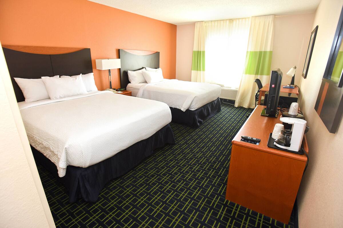 Quarto de hotel com duas camas, uma escrivaninha com computador e máquina de café, e uma janela com cortinas.