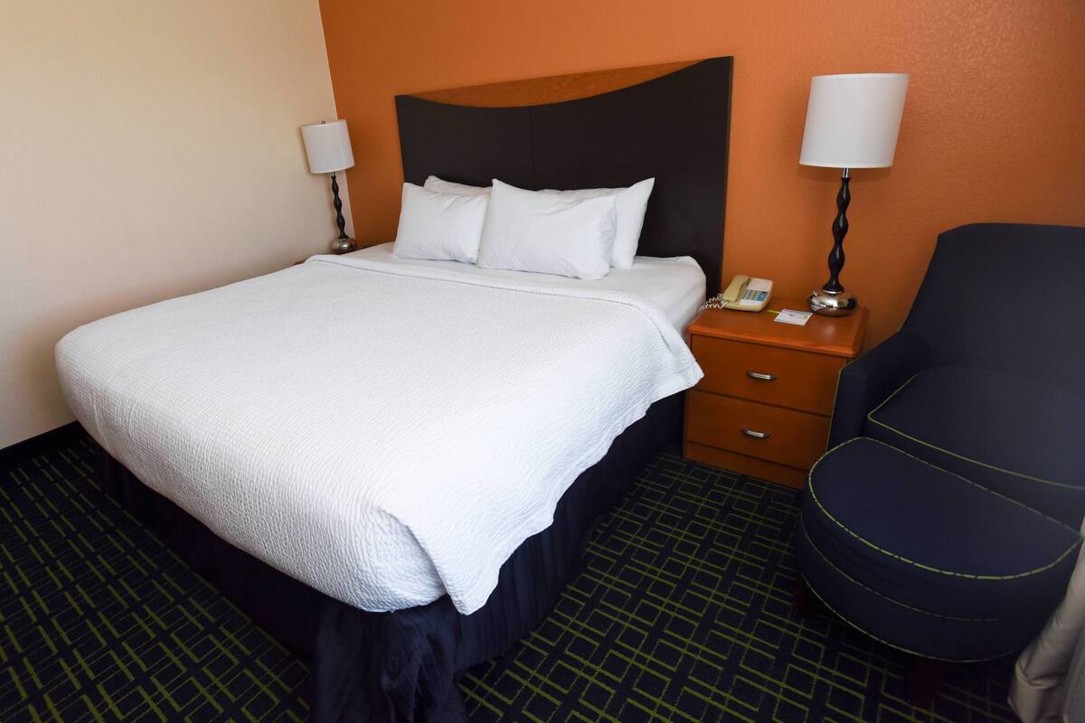 Quarto de hotel com uma cama, mesa de cabeceira com telefone e luminária, e uma cadeira.
