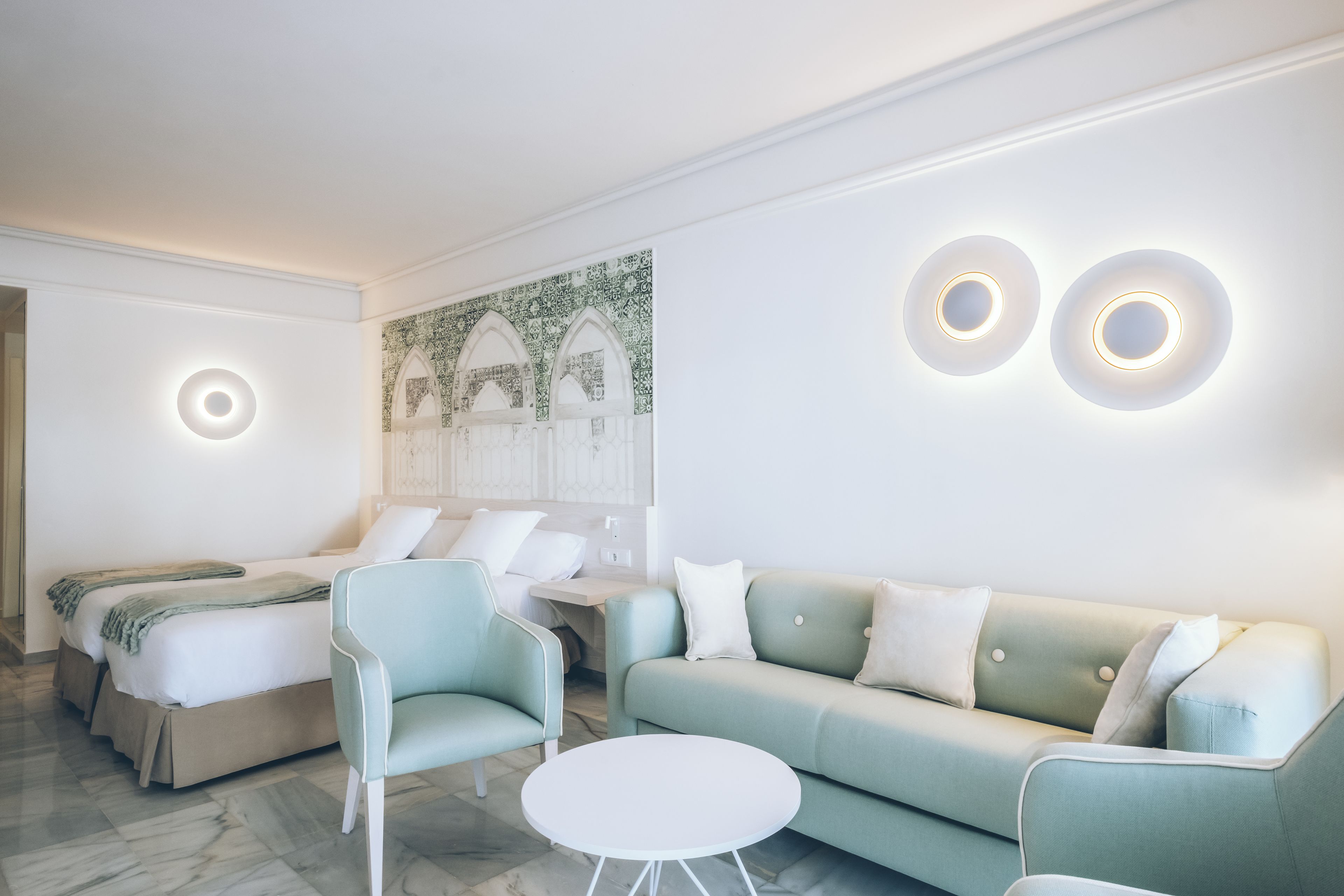 A modern hotel room with a bed, a light green sofa, a round white table, and two wall-mounted circular light fixtures.