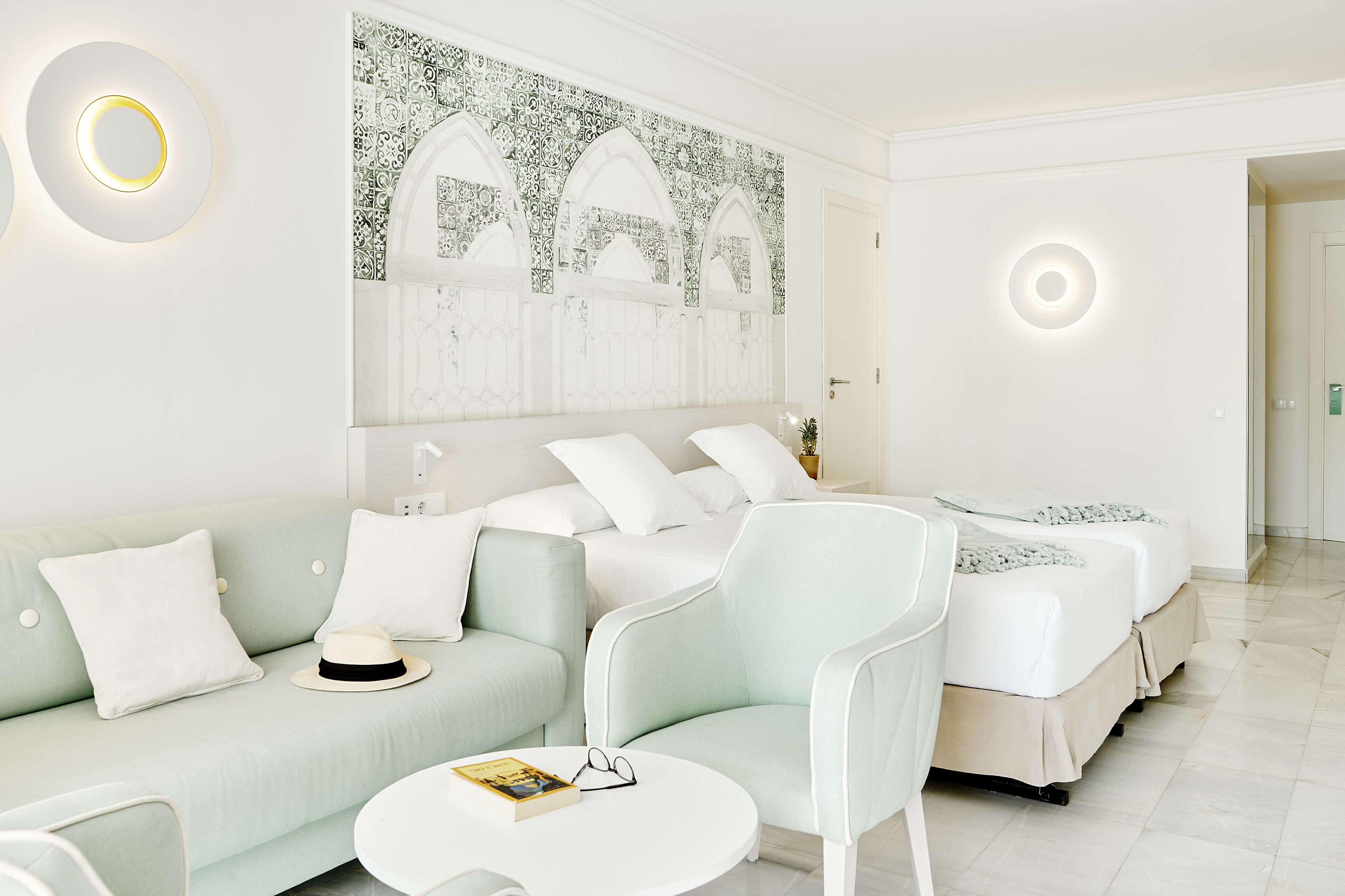 Une chambre d’hôtel avec un canapé vert clair, deux lits blancs, une table basse ronde et un mur orné d’un motif décoratif.