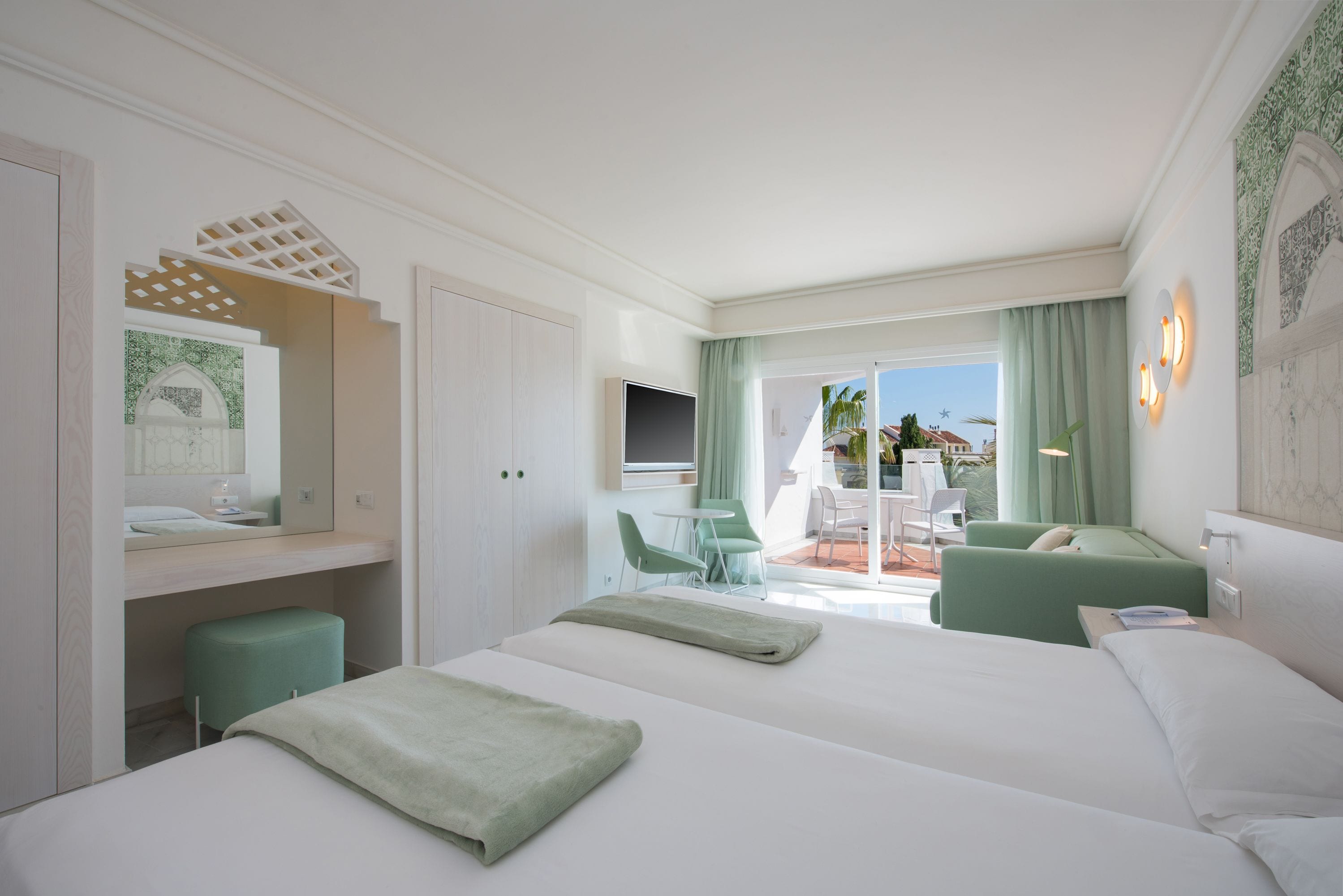 A hotel room with two beds, a TV, a balcony with a table and chairs, and a view of the city.