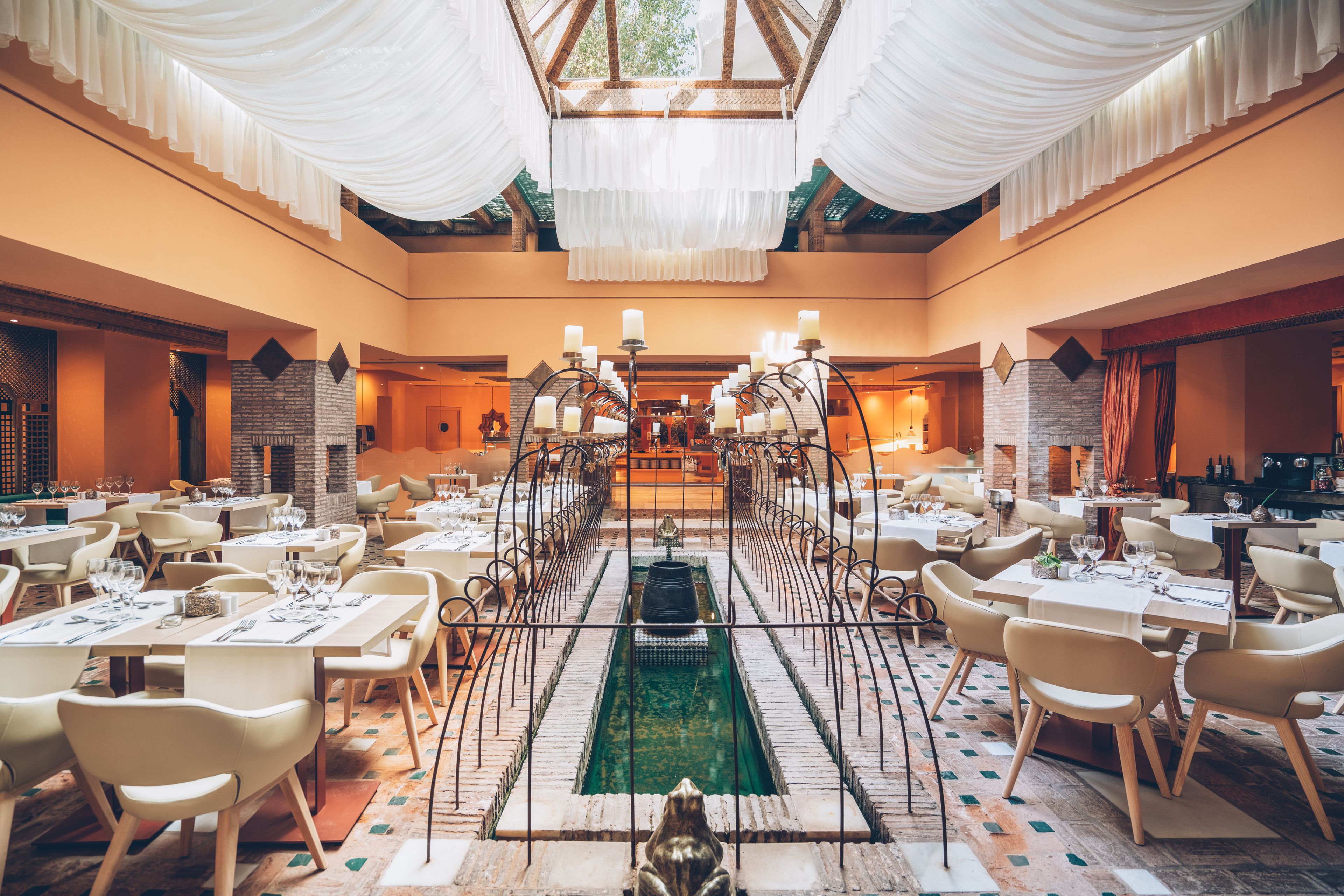 A spacious dining area with multiple tables and chairs, a central fountain, and a high ceiling with draped fabric.