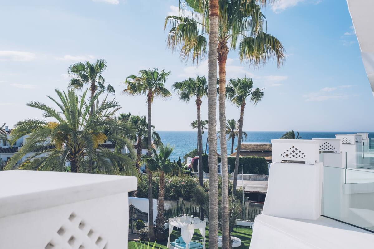 Un balcon avec des meubles blancs, des palmiers et une vue sur la mer.
