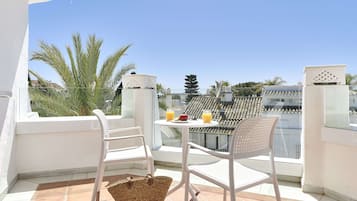 A balcony with white chairs and a table, a wicker basket, and a view of a cityscape with palm trees.