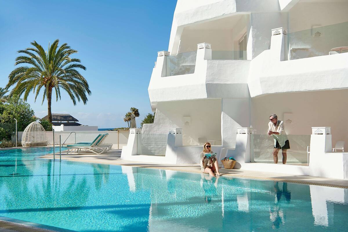 Un espace piscine avec une piscine à l’eau d’un bleu limpide, un bâtiment blanc doté de balcons et un palmier.
