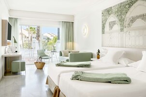 Hotelkamer met twee bedden, een bureau, een stoel en een balkon met uitzicht op palmbomen en gebouwen.