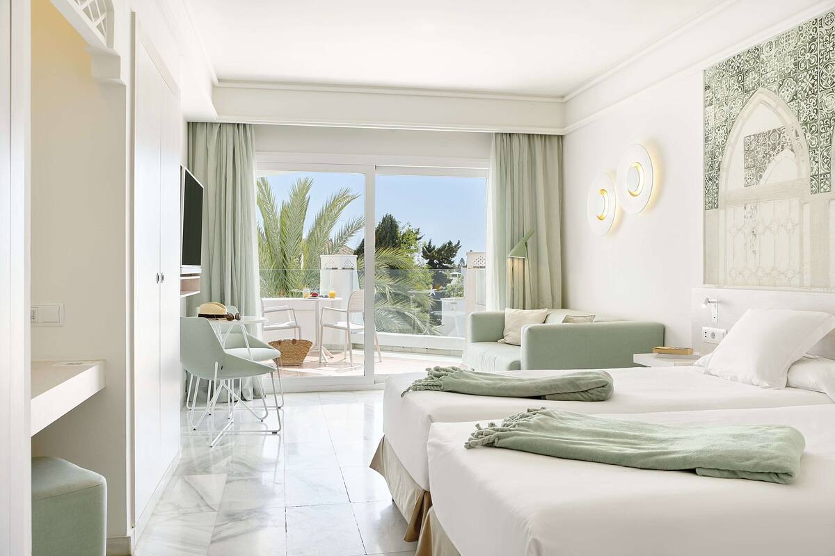 Une chambre d’hôtel avec deux lits, un bureau et un balcon donnant sur des palmiers.