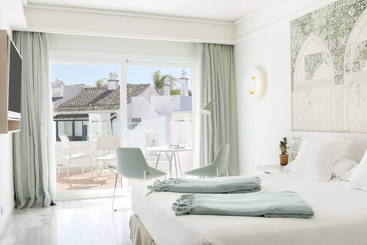 Une chambre d’hôtel avec un lit, deux chaises, une petite table, une plante en pot et une vue sur les bâtiments à travers une porte vitrée.
