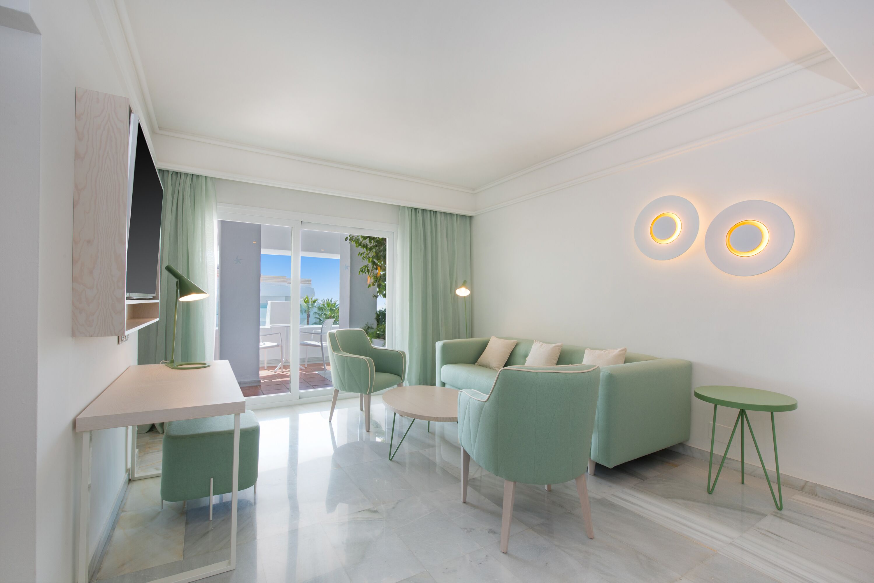 A modern living room with a green sofa, a small table, and a green chair. There is a window with curtains, a lamp, and two wall-mounted lights.