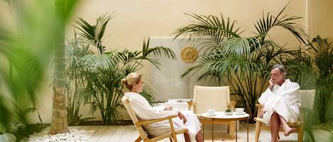 Two people in white robes seated at a round table with a cup and saucer, surrounded by tropical plants and a palm tree.