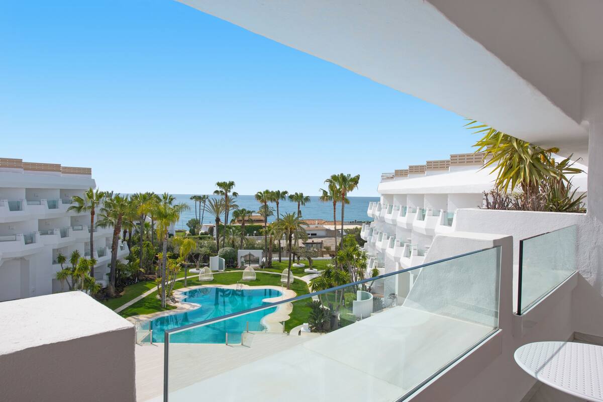 Un espace piscine offrant une vue dégagée sur l’océan, entouré de palmiers et bordé d’un bâtiment blanc doté de balcons.