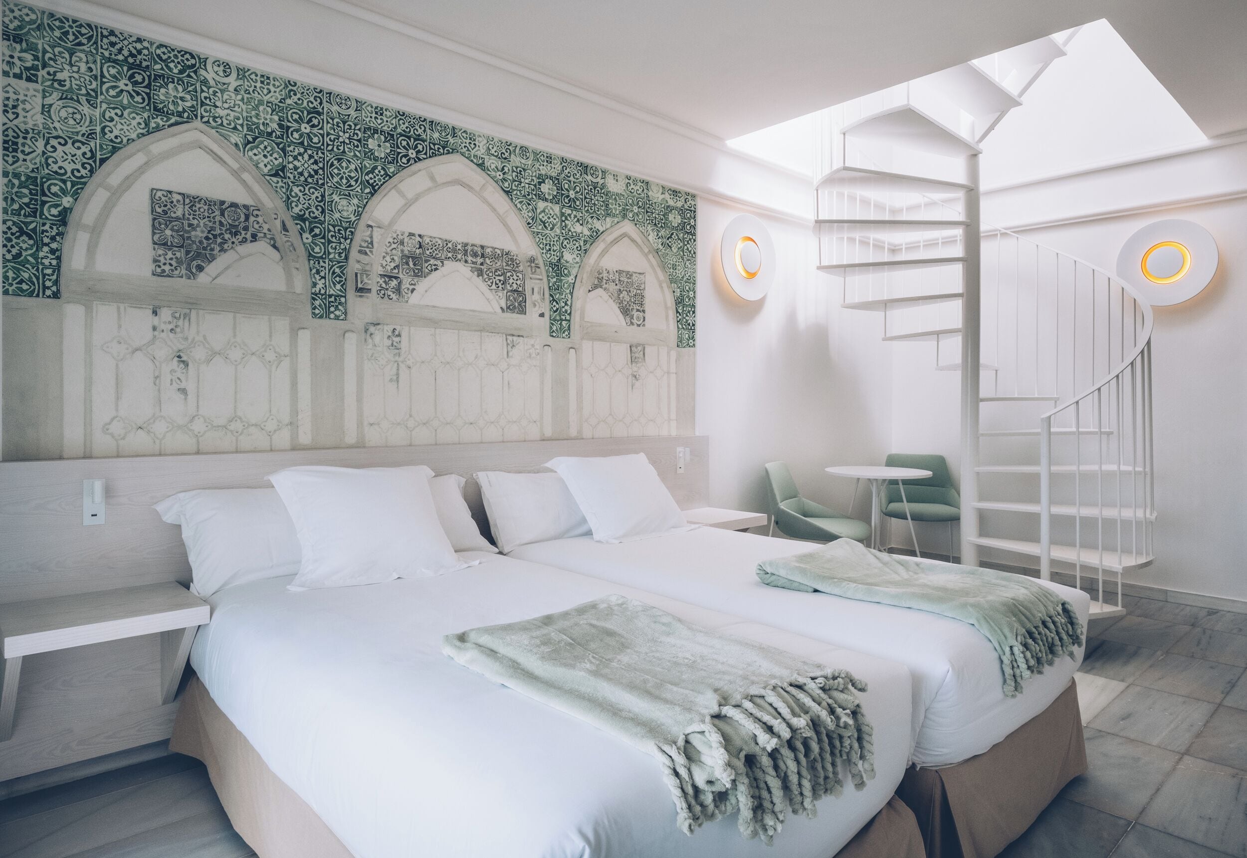 A modern hotel room with a large bed, a skylight, a spiral staircase, and a green chair.