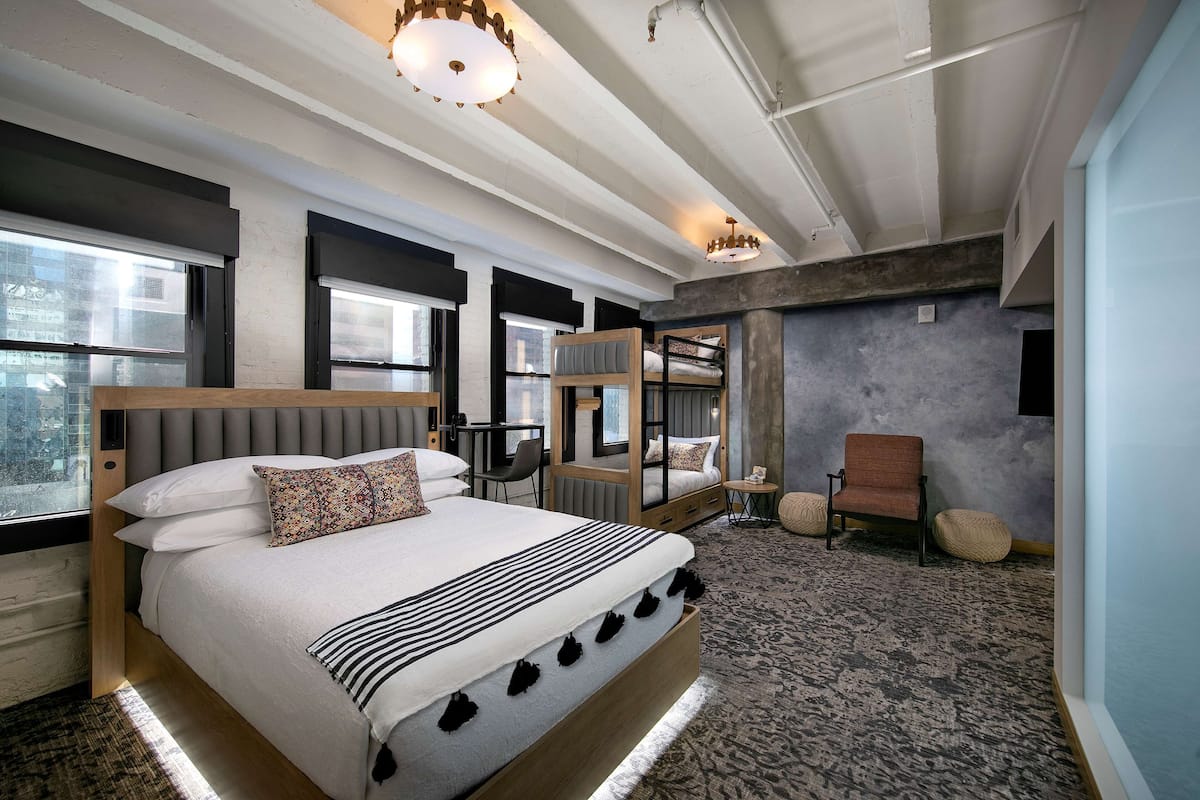 A modern hotel room with a large bed, a seating area with a chair and ottoman, a small table, and a view through large windows.