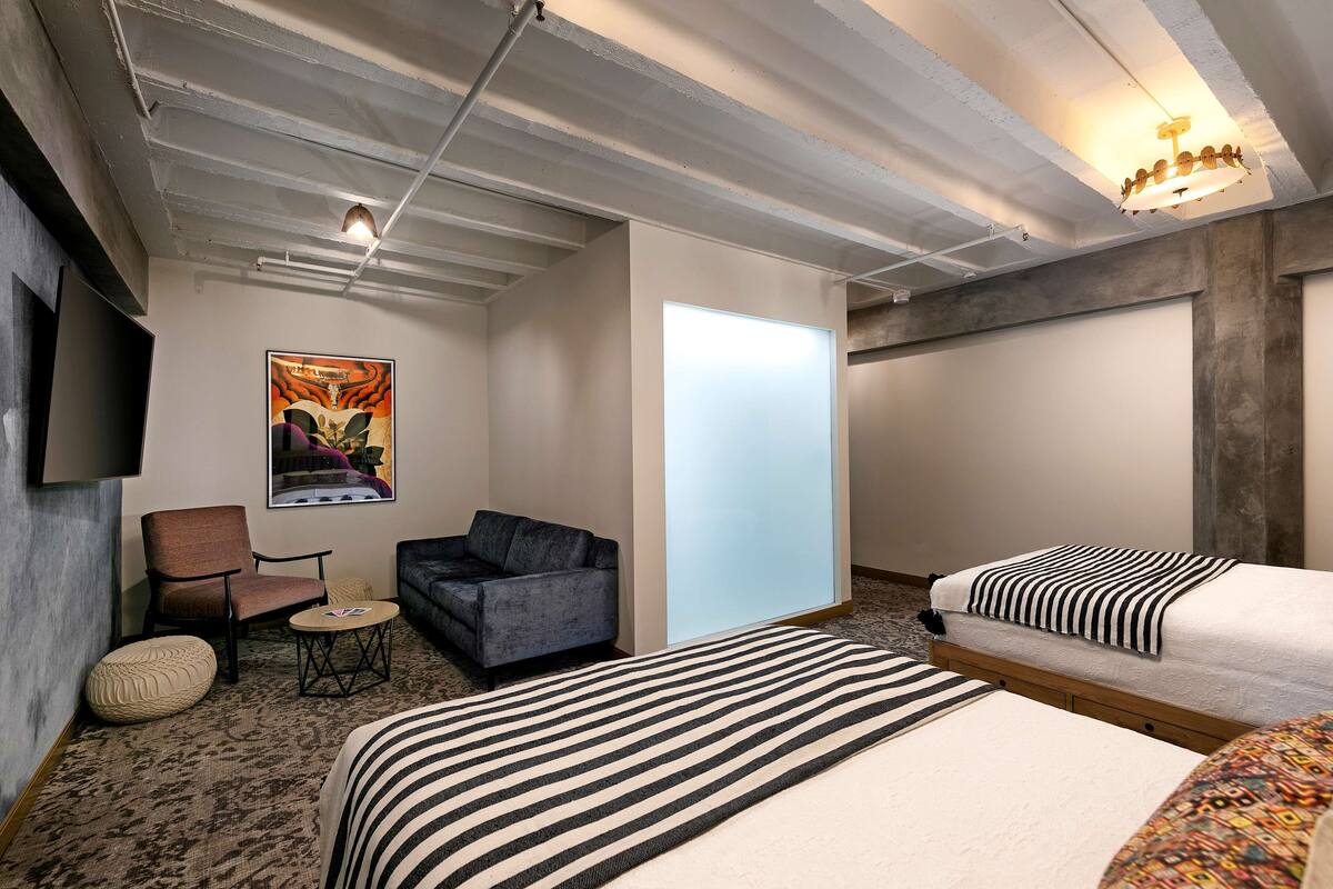 A hotel room with a bed, two sofas, a small table, a chair, a wall-mounted TV, and a ceiling light fixture.