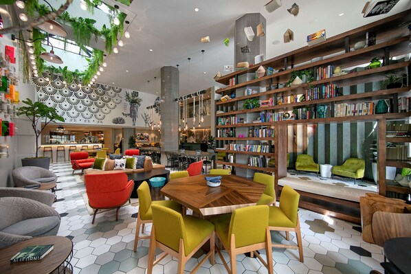 A modern indoor space with a wooden dining table surrounded by colorful chairs, a bookshelf filled with books, and a decorative wall with geometric patterns.