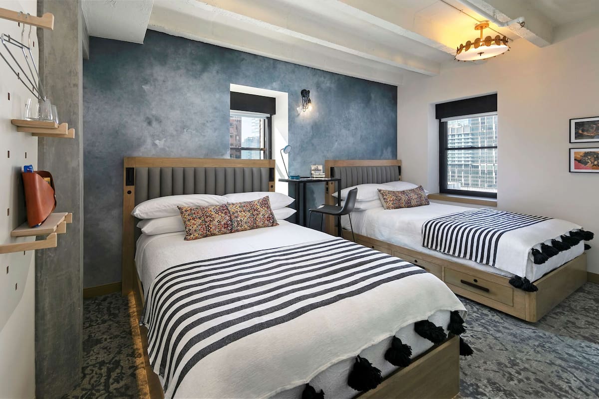 A modern hotel room with two beds, a small table, a chair, and a window with blinds.