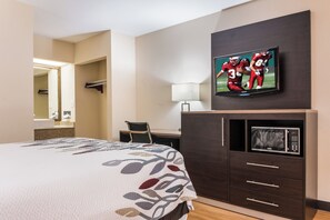 Habitación de hotel con cama, televisión montada en la pared, escritorio con silla, lámpara y microondas.