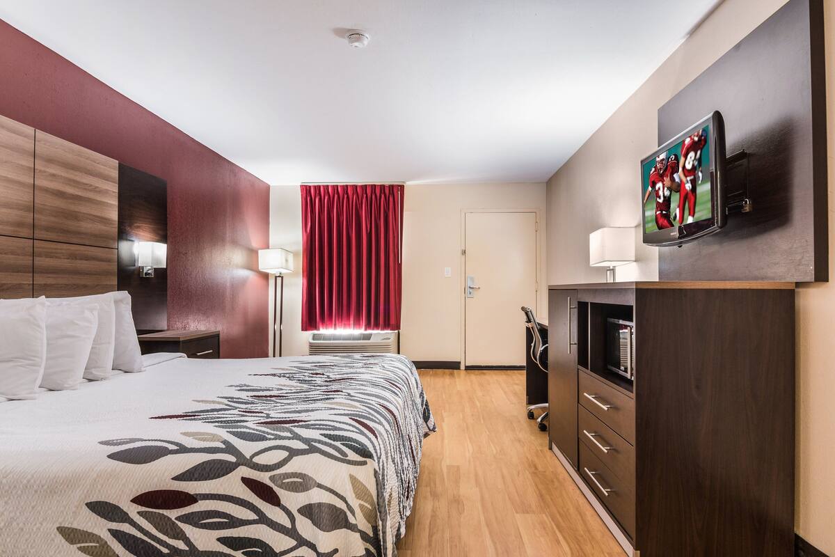 A hotel room with a bed, a TV mounted on the wall, a desk with a lamp, and a wardrobe.